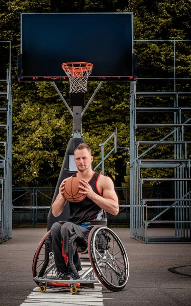 Joueur de basket-ball cramoisi dans un fauteuil roulant tient une balle sur un terrain de jeu ouvert . — Photo