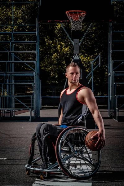 Joueur de basket-ball cramoisi dans un fauteuil roulant tient une balle sur un terrain de jeu ouvert . — Photo