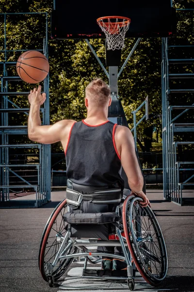 Visão traseira de um jogador de basquete com deficiência em cadeira de rodas girando basquete em seu dedo em campo de jogo aberto . — Fotografia de Stock