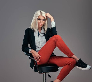 Sarışın kadın moda trendy kıyafetler ve stüdyoda bir sandalyede otururken poz gözlük. Gri bir arka plan üzerinde izole.