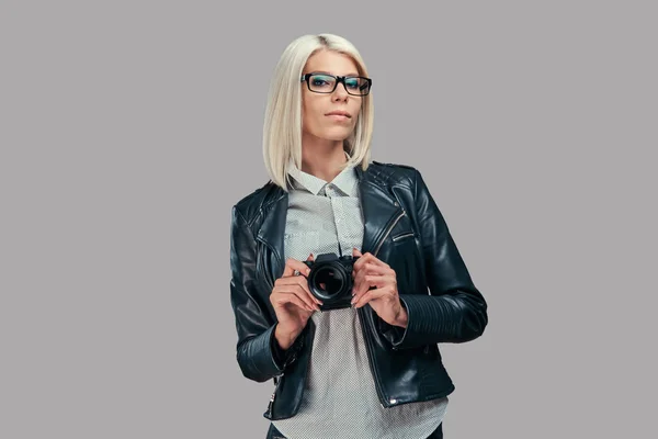 白いブラウスと黒の革ジャケットのスタジオでカメラにポーズをとってスマート ブロンド女性写真家の肖像画 灰色の背景に分離 — ストック写真