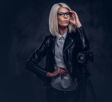 Trendy kıyafetler yaslanmış bir stüdyoda bir tripod ile profesyonel bir fotoğraf üzerinde görünüyor iken kameraya poz akıllı sarışın kadın fotoğrafçı.