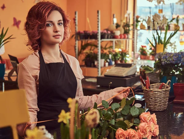 Чарівна руда жінка-флорист в уніформі робить красиву квіткову композицію, стоячи на стійці квіткового магазину . — стокове фото