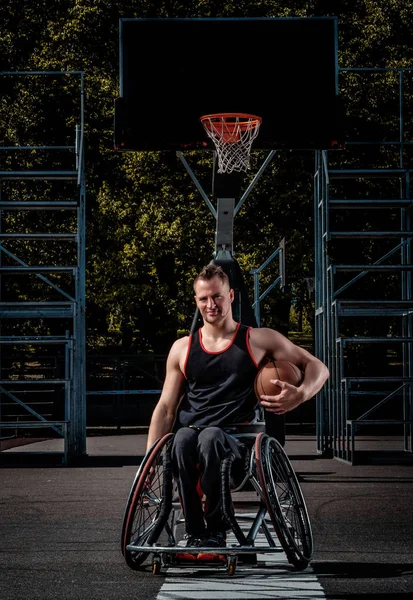 Un joueur de basket-ball handicapé souriant en fauteuil roulant tient une balle sur un terrain de jeu ouvert . — Photo