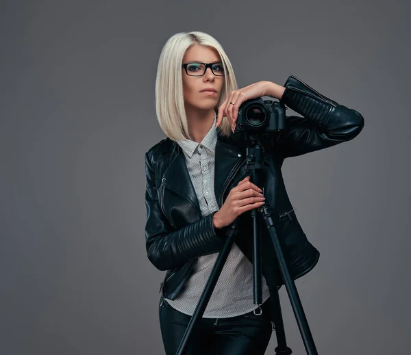 Fotógrafa loira inteligente em roupas da moda posando enquanto se inclina em uma câmera profissional com um tripé em um estúdio, olha para a câmera . — Fotografia de Stock