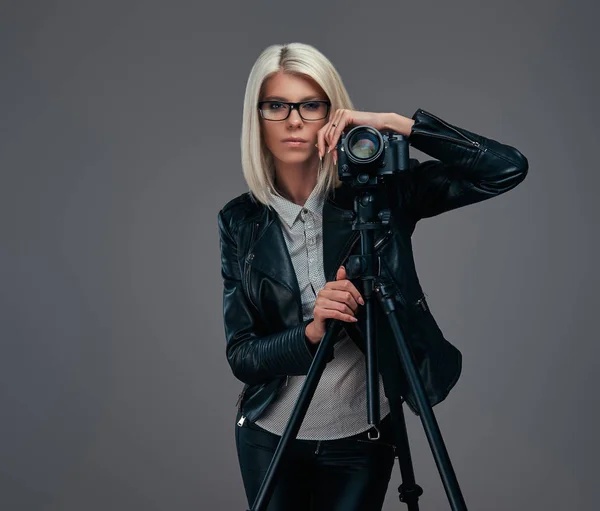 Smart blond kvinnlig fotograf i trendiga kläder poserar medan lutande på en professionell kamera med ett stativ på en studio, ser på kameran. — Stockfoto