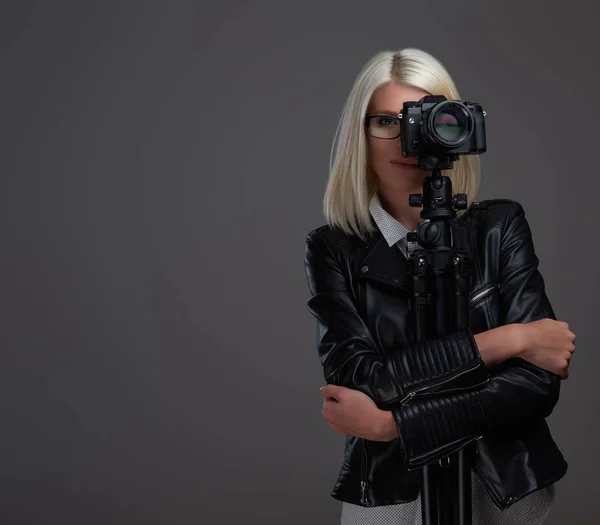 Beleza fotógrafa loira inteligente em óculos usando jaqueta de couro preto abraçando uma câmera profissional com um tripé enquanto tira uma foto em um estúdio . — Fotografia de Stock