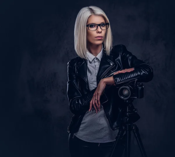 Intelligente fotografa bionda in abiti alla moda in posa mentre si appoggia su una fotocamera professionale con un treppiede in uno studio, guarda la fotocamera . — Foto Stock