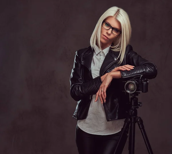 Chytrá blondýna fotografek v trendy oblečení, lodě, zatímco se opírá o profesionální kameru s stativu ve studiu, se dívá na kameru. — Stock fotografie