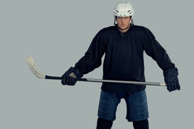 Hokey oyuncusu siyah koruyucu giysiler ve beyaz kask giymiş bir hokey sopası tutar. Gri bir arka plan üzerinde izole.