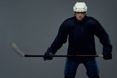 Hokey oyuncusu siyah koruyucu giysiler ve beyaz kask giymiş bir hokey sopası tutar. Gri bir arka plan üzerinde izole.