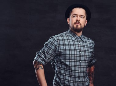 Portre sakal ve saç modeli ile dövmeli orta yaş hipster birinin bir kareli gömlek ve şapka giymiş, bir stüdyoda poz. Karanlık dokulu bir arka plan üzerinde izole.