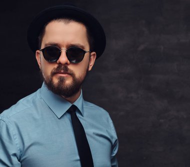 Portre sakal ve saç modeli ile yakışıklı bir orta yaşlı adamın şapka ve güneş gözlüğü ile resmi elbise giymiş. Bir stüdyoda dokulu koyu arka plan üzerinde izole.