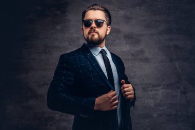 Giyinmiş emin yakışıklı şık orta yaşlı adam sakal ve saç modeli ile zarif bir resmi takım elbise ve Studio dokulu koyu arka plan üzerinde güneş gözlüğü.
