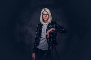 Akıllı sarışın kadın fotoğrafçı profesyonel bir kamera ile bir stüdyoda bir tripod üzerinde bir eğilimi görünüyor iken kameraya poz trendy kıyafetler. Karanlık bir arka plan üzerinde izole.