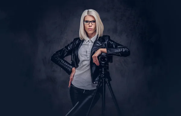 穿着时髦衣服的金发女摄影师 在摄影棚里靠着一个三脚架的专业相机 看着相机 在黑暗背景下被隔离 — 图库照片