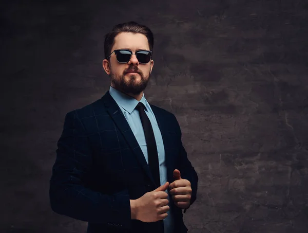 Confident handsome fashionable middle-aged man with beard and hairstyle dressed in an elegant formal suit and sunglasses on a textured dark background in studio.