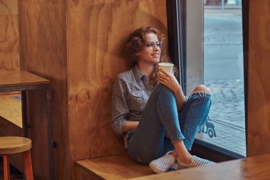 Rahat kıyafetler ve paket servisi olan restoran kahve ile bir pencere pervazına oturan gözlük gülümseyen genç redhead kıvırcık kız.