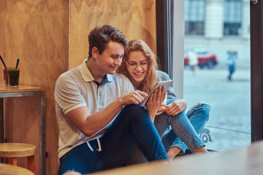 İki mutlu bir genç öğrenciler üniversite kantin, bir pencere pervazına üzerinde oturan bir mola sırasında dijital tablet kullanma.