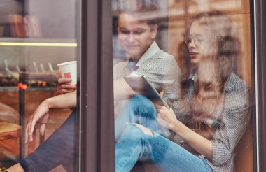 Kahve içme ve dijital tablet bir mola sırasında bir üniversite kampüsünde bir pencere pervazına üzerinde otururken kullanma iki dalgın genç öğrenciler.