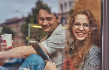 Mutlu genç öğrencilerin kahve içme ve pencere pervazına bir üniversite kampüsünde bir mola sırasında oturan dijital tablet kullanma.