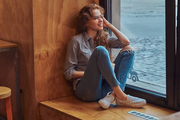 カジュアルな服やテイクアウトのコーヒーを飲みながら窓の土台の上に座ってメガネを着て笑顔の若い赤毛巻き毛の少女 — ストック写真