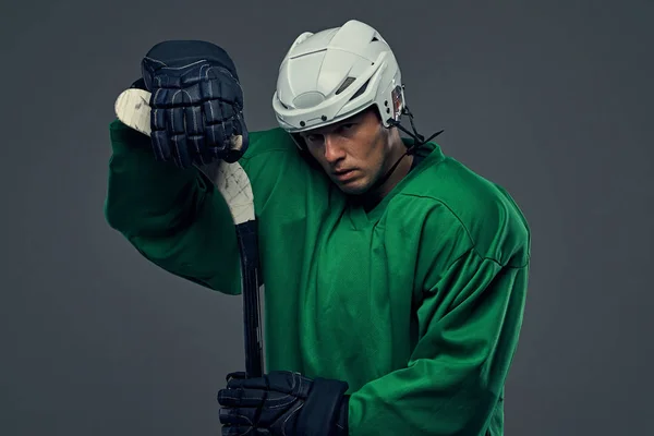灰色の背景に緑色の防護服と白いヘルメット立ってアイス ホッケー用スティックを着て疲れホッケー選手 — ストック写真