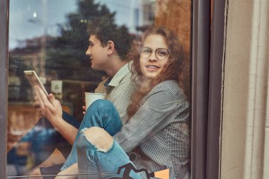 Mutlu genç öğrencilerin kahve içme ve pencere pervazına bir üniversite kampüsünde bir mola sırasında oturan dijital tablet kullanma.