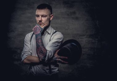 Beyaz gömlek ve pantolon askısı giyen düşünceli bir eski moda dövmeli adam portresi bir silindir şapka ve el çene üzerinde tutar. Karanlık bir arka plan üzerinde izole.