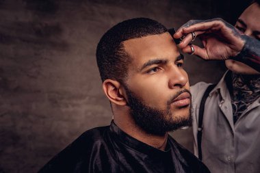 Bearded African American hipster guy getting a haircut by an old-fashioned tattooed professional hairdresser does the haircut. clipart