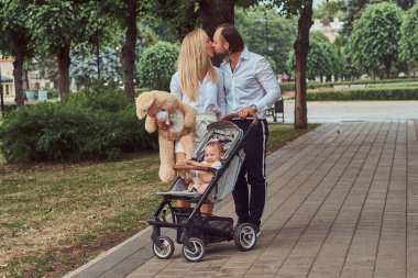 Mutlu genç Aile kavramı. Güzel sarışın anne ve bebek arabası içinde onların küçük kızı ile parkta yürüyüş sakallı şık baba.