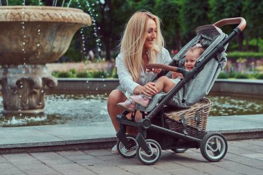 Mutlu sarışın kadın beyaz bir bluz ve bir bebek arabası bir çeşme şehir Parkı yakınındaki küçük kızıyla birlikte poz şort giymiş.