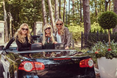 Tatil ve insanlar kavramı. Üç mutlu sarışın bayan arkadaşlar tatil yaz cabriolet arabada oturan güneş gözlüğü moda giysiler giyiyor.
