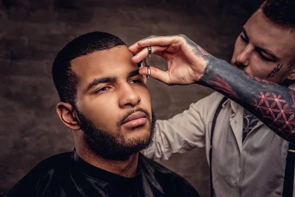 老式的专业纹身理发师用剪刀和梳子给非洲裔美国人的客户理发 在深色纹理背景下隔离 — 图库照片