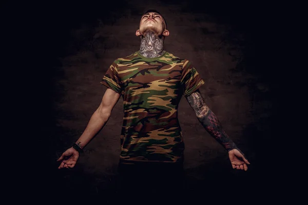 Tattooed young guy in a military t-shirt pose at the studio. Isolated on a dark textured background.