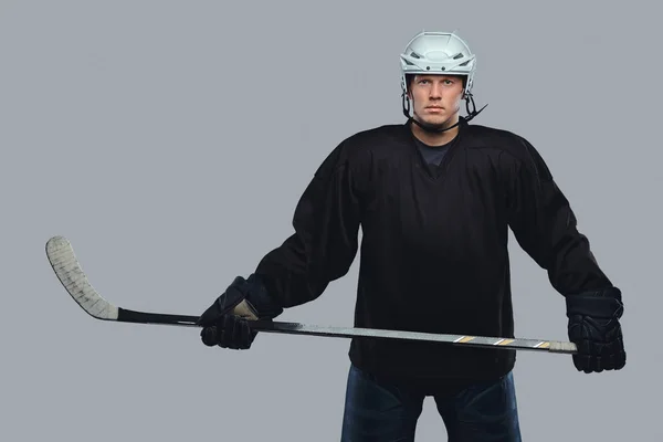 Hockey player wearing black protective gear and white helmet holds a hockey stick. Isolated on a gray background.