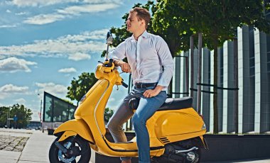 Beyaz bir gömlek ve bir gökdelen karşı sarı klasik İtalyan scooter üzerinde oturan kot giymiş şık saç kesimi ile kendinden emin yakışıklı adam. 