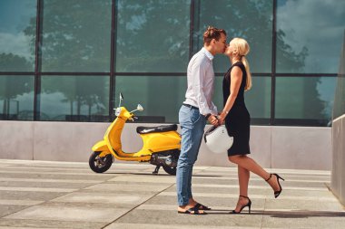 Aşık - şık yakışıklı beyaz tişört ve kot pantolon ve siyah elbise giyen sevimli sarışın kadın giymiş güzel çift el ele tutuşup karşı bir gökdelen öp.