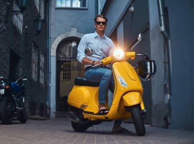 Genç adam şık giyinmiş bir beyaz bir gömlek ve pantolon akşam eski bir Europe street üzerinde sarı klasik İtalyan scooter üzerinde binmek.