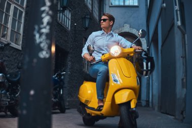 Genç adam şık giyinmiş bir beyaz bir gömlek ve pantolon akşam eski bir Europe street üzerinde sarı klasik İtalyan scooter üzerinde binmek.