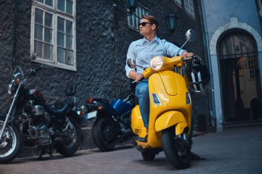 Genç adam şık giyinmiş bir beyaz bir gömlek ve pantolon akşam eski bir Europe street üzerinde sarı klasik İtalyan scooter üzerinde binmek.