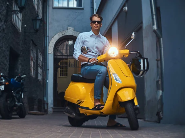Genç adam şık giyinmiş bir beyaz bir gömlek ve pantolon akşam eski bir Europe street üzerinde sarı klasik İtalyan scooter üzerinde binmek.