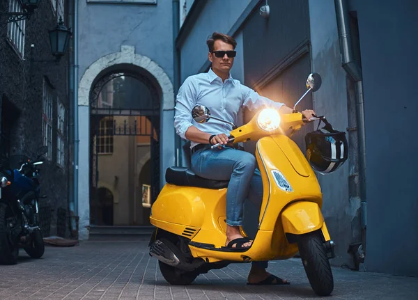 Young stylish guy dressed in a in a white shirt and jeans ride on yellow classic italian scooter on an old Europe street in the evening.