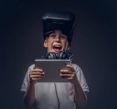 Sürpriz bir küçük çocuk portresi sanal gerçeklik gözlüklü beyaz tişört giymiş ve kulaklık dijital tablet koyu arka plan üzerinde izole bir stüdyoda holding. 