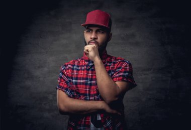 Dalgın bir Afrikalı-Amerikalı hipster adam kırmızı Polar gömlek ve cap adlı stüdyo giymiş. Karanlık dokulu bir arka plan üzerinde izole.