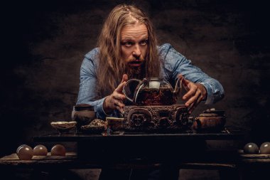 Çay töreni kavramı. Mavi gömlekli, çay, el yapımı bir çay seti kullanarak yapmaya konsantreleri portre bir kızıl saçlı hippi erkek uzun saç ve tam sakal ile giyinmiş. Karanlık dokulu bir izole