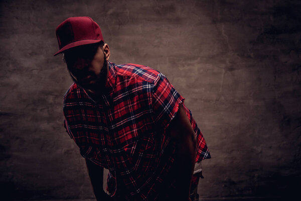Portrait of an African-American hipster guy dressed in a red fleece shirt and cap at the studio. Isolated on a dark textured background.
