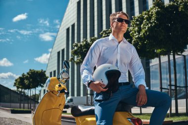 Emin yakışıklı şık saç kesimi ile beyaz bir gömlek, kot pantolon ve güneş gözlüğü tutar kask ve bir gökdelen karşı sarı klasik İtalyan scooter üzerinde oturan giymiş.