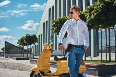 Kendine güvenen yakışıklı şık saç kesimi ile beyaz bir gömlek giymiş, kot pantolon ve güneş gözlüğü tutan sarı klasik İtalyan scooter yakınındaki bir kask ayakta karşı bir gökdelen.
