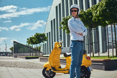 Beyaz bir gömlek, kot pantolon, güneş gözlüğü ve motosiklet kask ayakta ile içinde şık saç kesimi ile kendinden emin yakışıklı adam silah sarı klasik İtalyan scooter bir gökdelen karşı yakınındaki geçti.